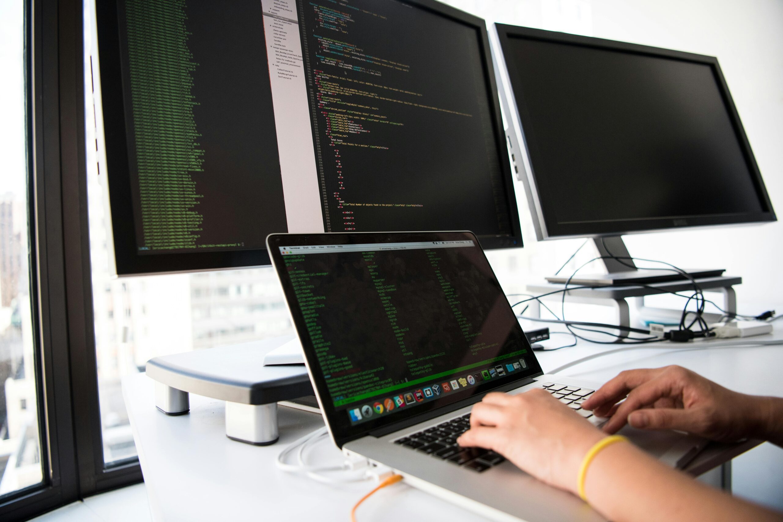 Développeur web et web mobile A developer writes code on a laptop in front of multiple monitors in an office setting.